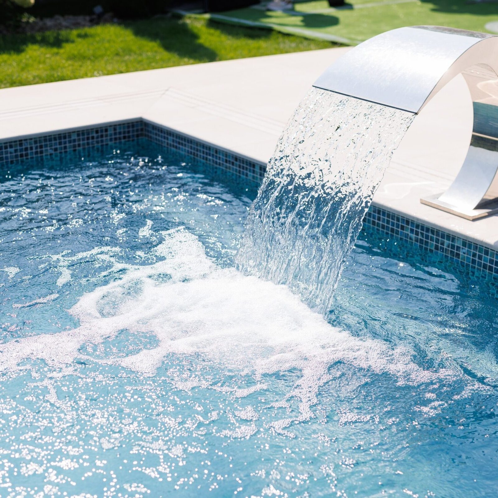 A,Modern,Swimming,Pool,With,A,Sleek,Water,Feature,Pouring A modern swimming pool with a sleek water feature pouring water into the pool. The water is clear and blue, creating a refreshing atmosphere. The surrounding area is well-maintained with green grass.